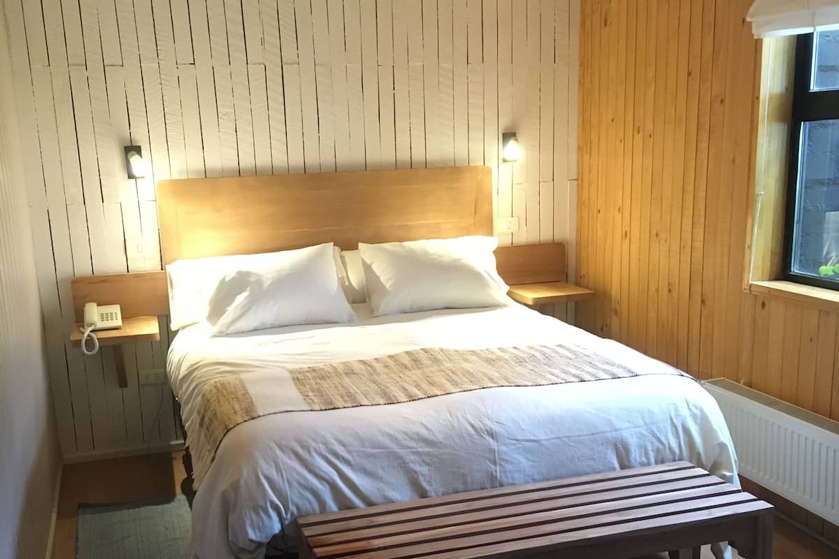 A bedroom with a wooden headboard, a bed with white linens, a bedside table with a lamp, a bench, and a patterned rug.