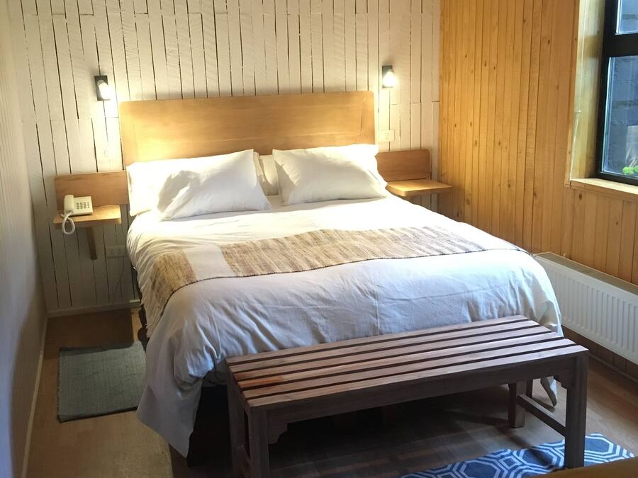 Un dormitorio con cabecera de madera, cama con ropa de cama blanca, mesita de noche con lámpara, un banco y una alfombra estampada.