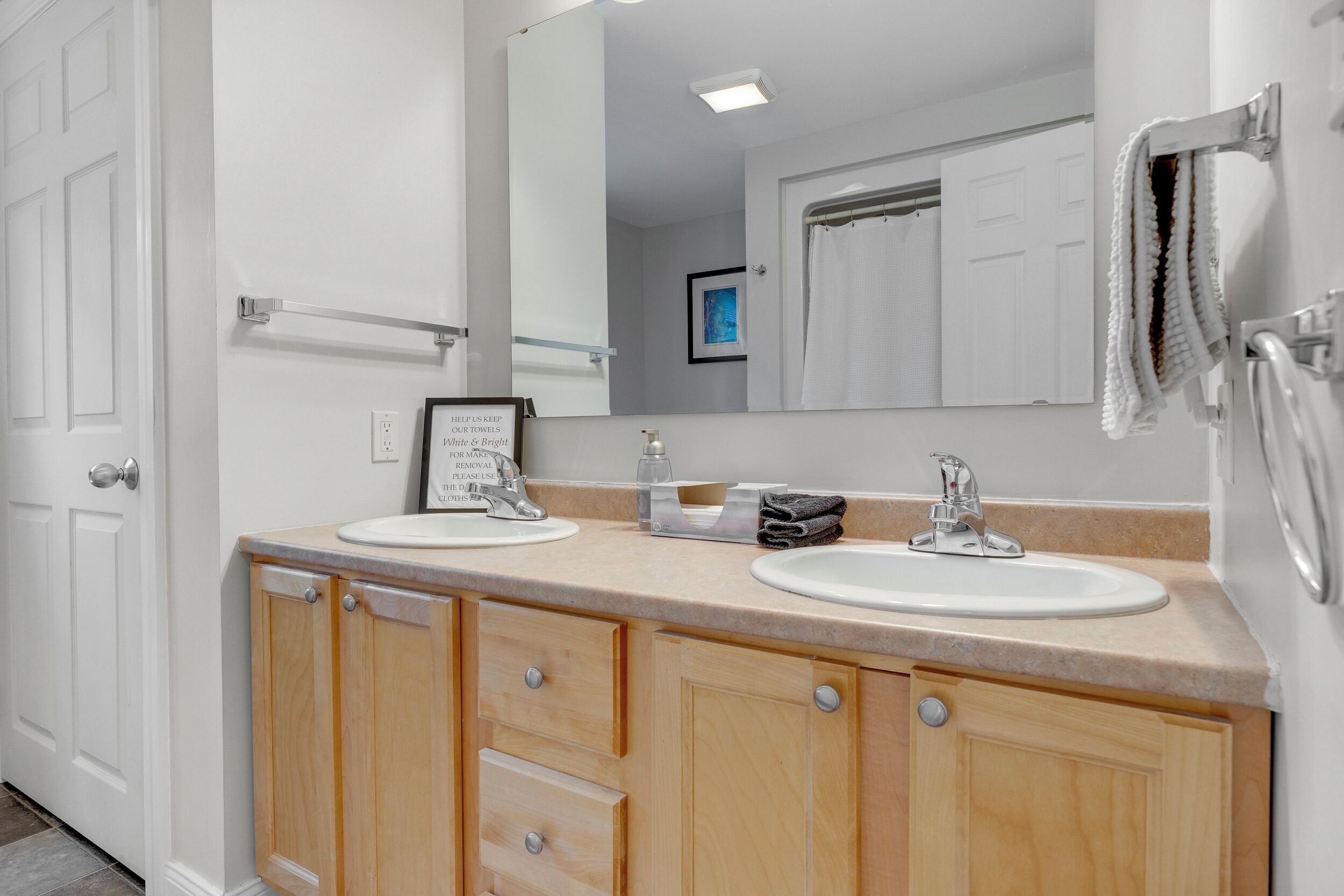 A bathroom with two sinks, a mirror, a towel rack, and a framed sign.