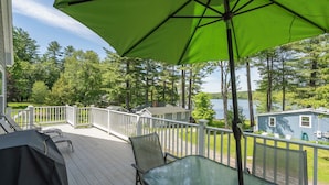 A deck with a table and chairs, a green umbrella, and a view of a lake and trees. - Lakefront haven near Pleasant Mt-central location-25 mi. to N.Conway (Bridgton)