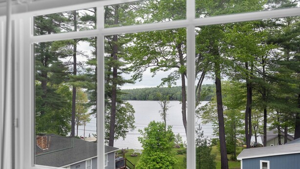 A view through a window of a lake surrounded by trees and a small house. - Lakefront haven near Pleasant Mt-central location-25 mi. to N.Conway (Bridgton)