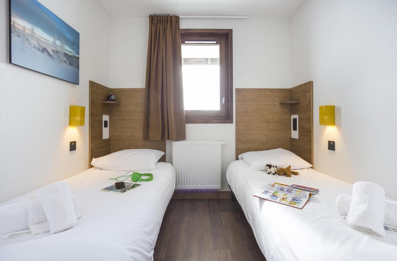 A hotel room with two single beds, a window with brown curtains, a radiator, and two wall-mounted light fixtures.