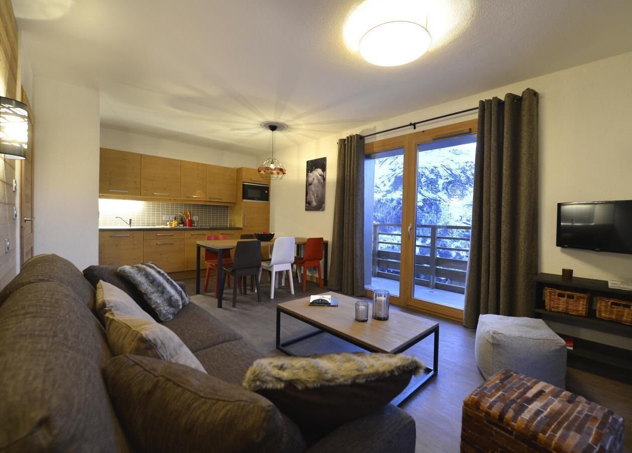 A living room with a sofa, ottoman, coffee table, and a TV. There is a dining area with chairs and a table, and a kitchen in the background.