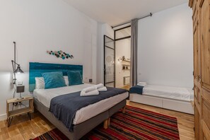 A modern bedroom with a large bed, a smaller bed, a nightstand, a lamp, a mirror, and a wooden wardrobe.