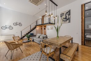 A modern interior with a wooden dining table, chairs, a staircase, and a living area with a sofa and cushions.