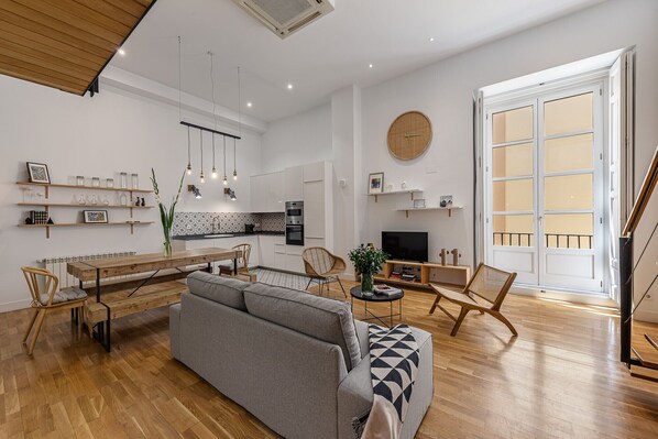 A modern living space with a dining area, sofa, TV, and kitchen.