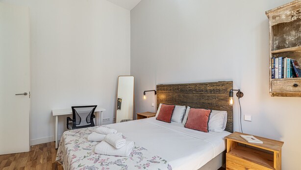 Un dormitorio con cabecera de madera, cama con ropa de cama floral, mesita de noche, una silla y un estante con libros.