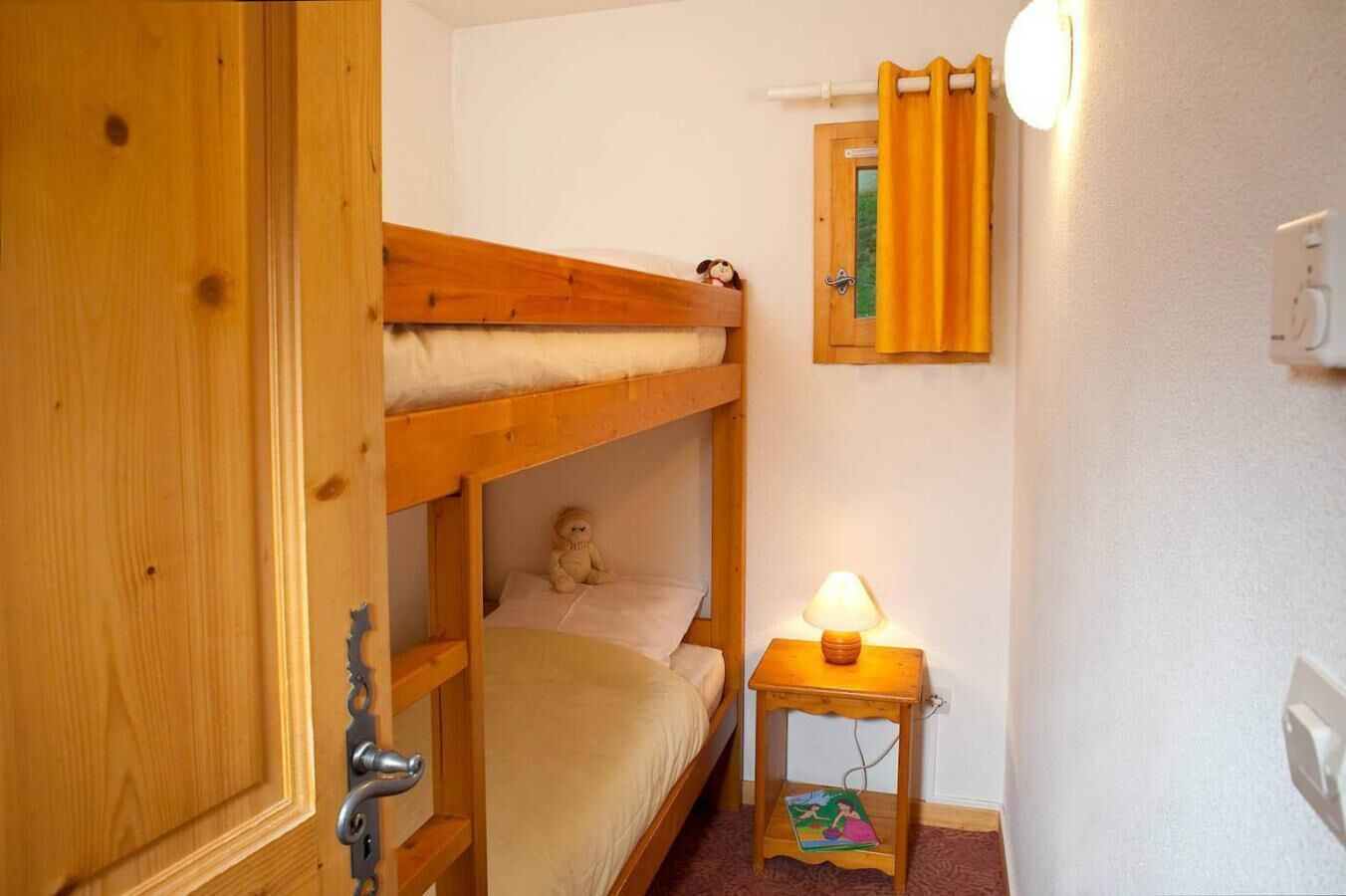 A bunk bed room with a wooden door, a bedside lamp, and a small table with a book.