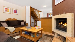 Un salon chaleureux avec un canapé, une table basse en bois sur laquelle sont posés des livres et un panier de fruits, une cheminée et un escalier menant à un autre niveau.
