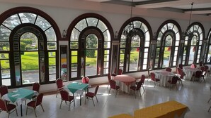 Uma ampla área de refeições interna, com várias mesas e cadeiras, grandes janelas em arco e vista para uma paisagem verde.