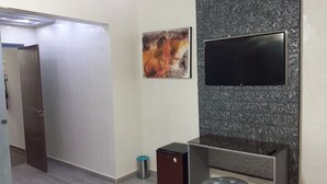 Quarto moderno com televisão de tela plana presa em uma parede texturizada, uma lixeira pequena, um armário de madeira escura e um banquinho acolchoado.