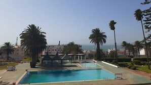 Uma piscina com três gazebos, palmeiras e vista para o mar.