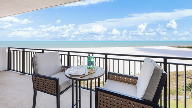 A balcony with two chairs and a small table, overlooking a beach with clear skies and calm waters. - Modern 11th Floor Beachfront Condo | Views/Pool (Marco Island)