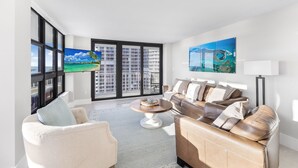 Una sala de estar moderna con un sofá color beige, un sillón blanco y una mesa de centro. Amplias ventanas ofrecen vistas a un paisaje urbano y palmeras.