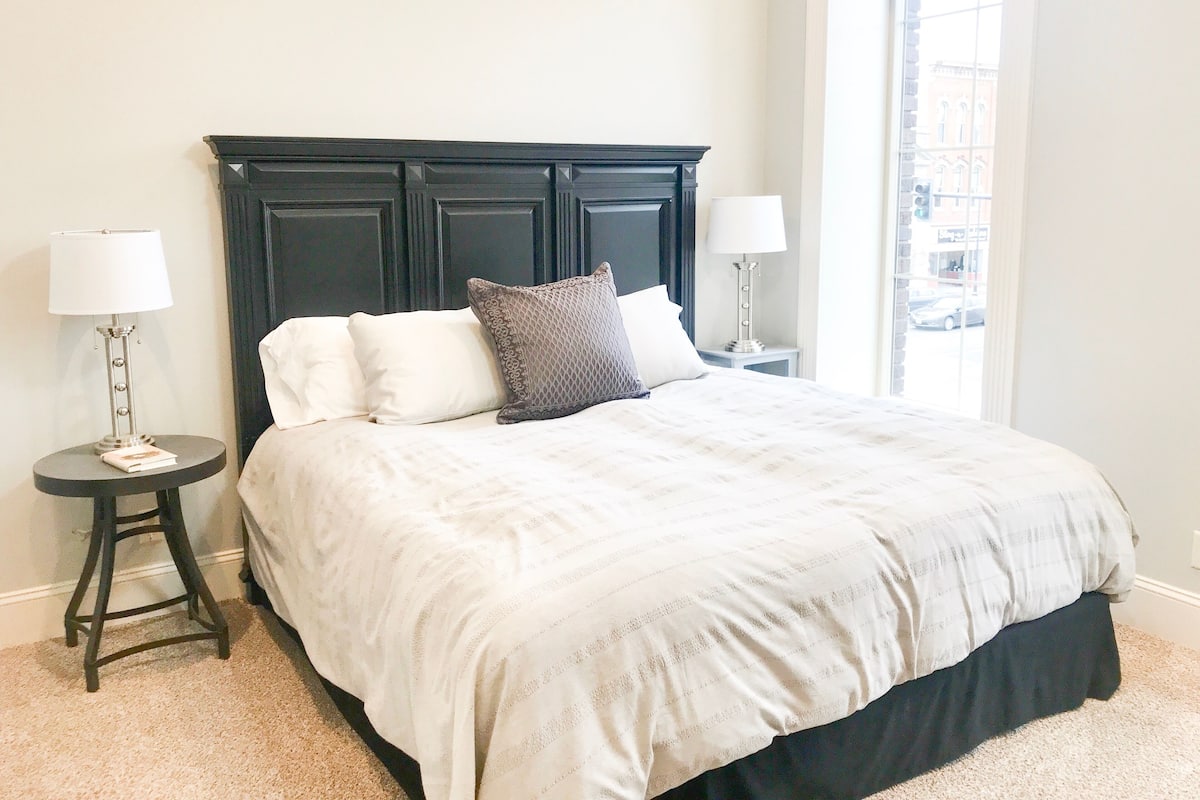A neatly made bed with a dark headboard, two bedside tables with lamps, and a window with a view of buildings.