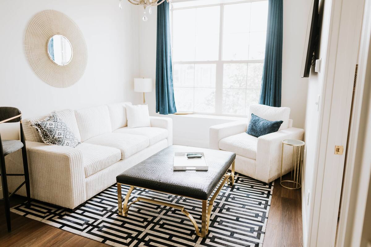 A modern living room with a white sofa, a black ottoman, and a patterned rug.