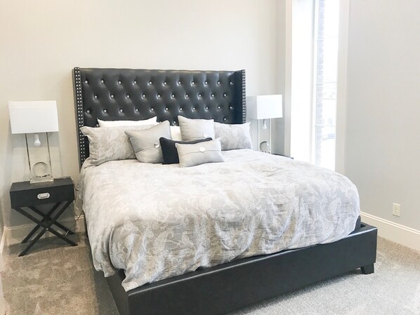 Une chambre moderne avec une tête de lit capitonnée, un grand lit recouvert d’une couvre-lit à motifs, deux tables de chevet avec des lampes et une vue sur la ville par la fenêtre.