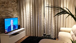A modern living room with a flat-screen TV on a white console, beige curtains, a black rug, and a beige sofa. - 2 double  Bedrooms  2 Bath Apartment With A/C Bugibba close to the sea  (St Paul's Bay)