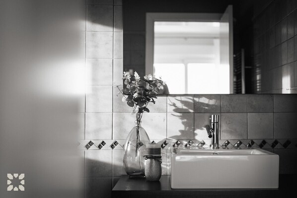 Un baño con un lavamanos blanco, un florero con flores y un espejo sobre el lavamanos.