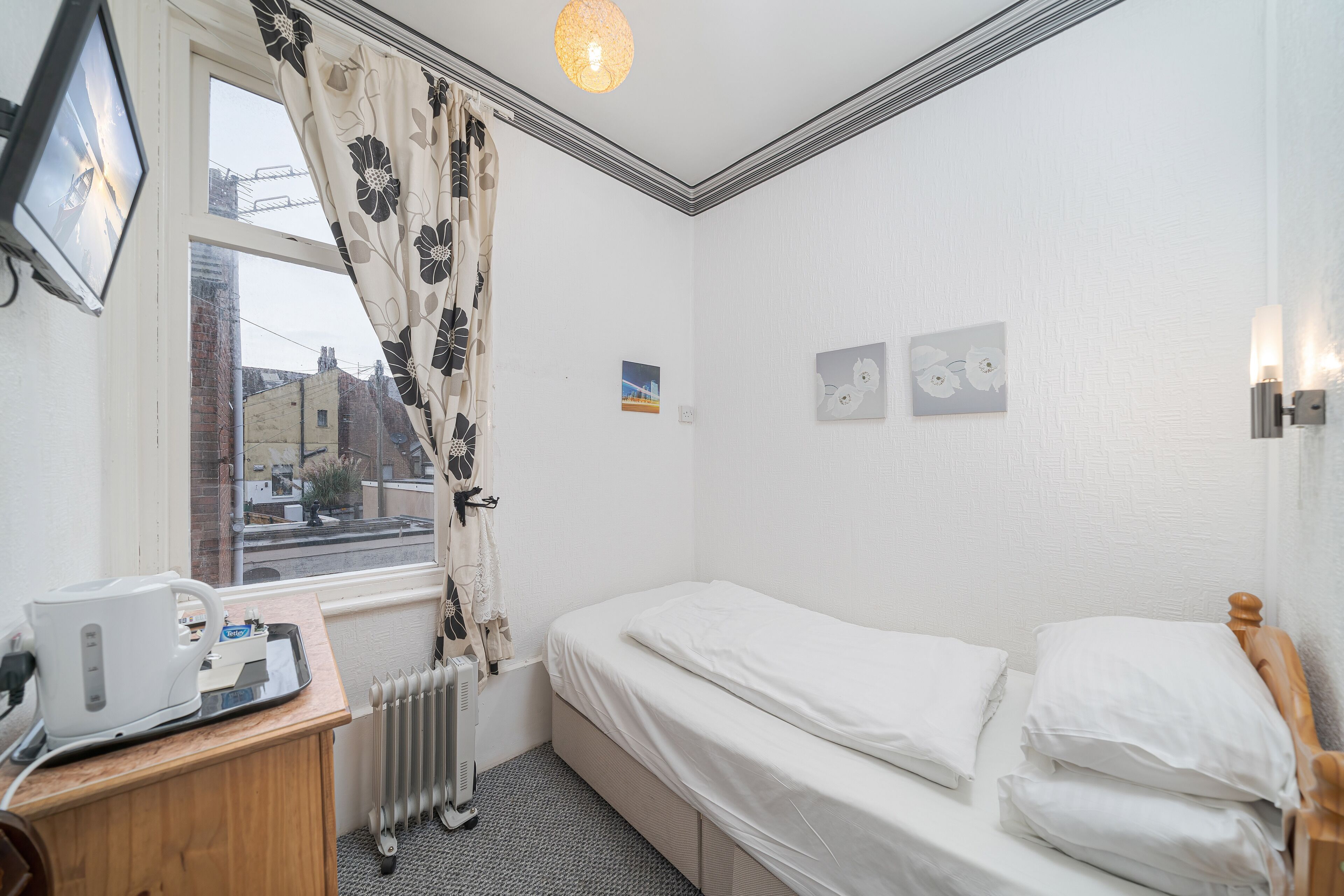 A small, clean room with a single bed, a radiator, a window with curtains, a small table with a kettle, and a picture on the wall.