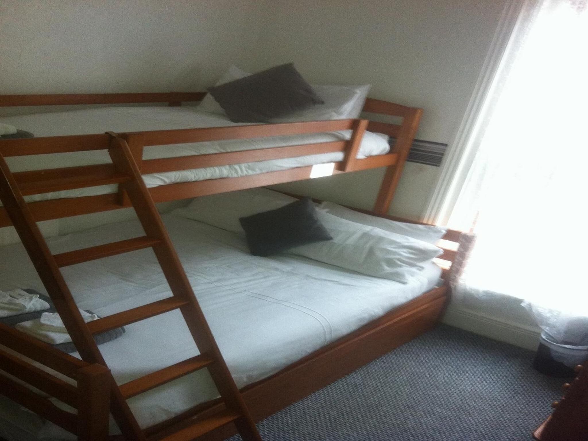 Uma cama de beliche com escada de madeira, roupa de cama branca e uma pequena lixeira.
