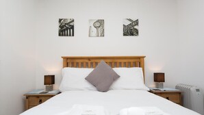 A neatly made bed with white linens and two pillows, flanked by bedside tables with lamps. Above the bed, there are three framed pictures on the wall. - Deneside Guest House (Blackpool)
