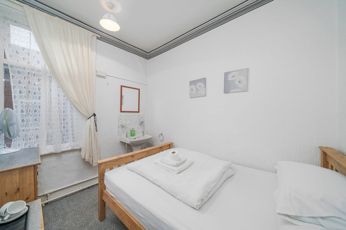 A clean, well-lit room with a bed, a wooden nightstand, a small table with a fan, and a bathroom with a sink and mirror.