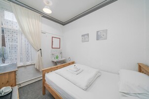 Una habitación limpia y bien iluminada con una cama, una mesita de noche de madera, una pequeña mesa con un ventilador y un baño con lavabo y espejo.