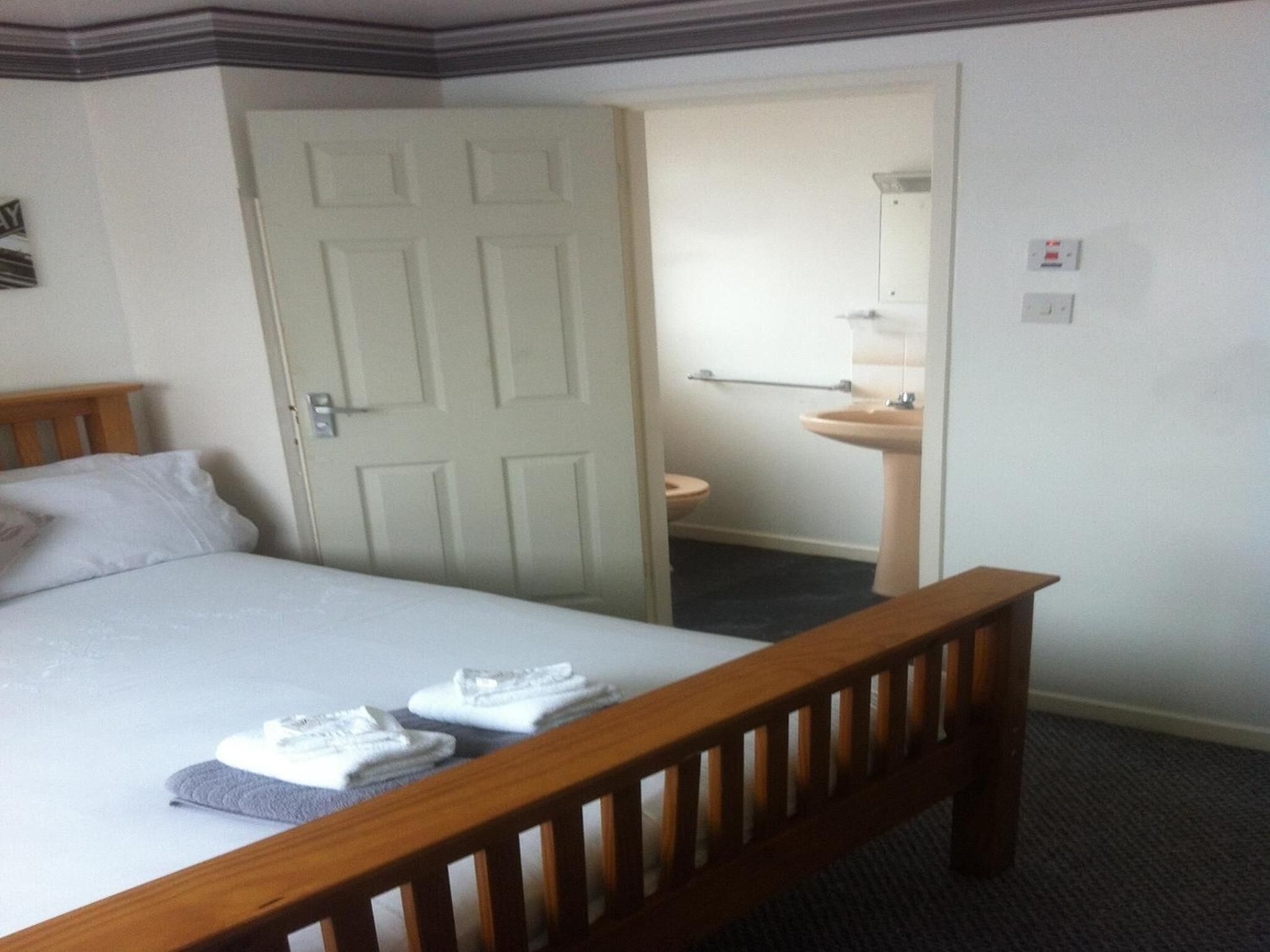A hotel room with a bed, a wooden headboard, a door, a bathroom with a sink and mirror, and a carpeted floor.