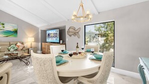 Uma sala de jantar com uma mesa redonda, cadeiras brancas e um lustre. Há uma televisão na parede exibindo "Roevens Vacations".
