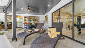 Un espacio de estar al aire libre techado, con sillones de mimbre, ventilador de techo y puerta de cristal que da a un ambiente interior.