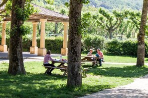 Ein überdachter Pavillon mit Picknicktischen in einem Park mit Bäumen und Blick auf Hügel.
