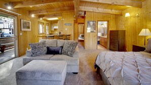A cozy log cabin-style room with a sofa, ottoman, and a bed. There is a kitchen area with a dining table and chairs, and a sign indicating the direction to the lodge. - Switzerland SKI Chalet #845 (Big Bear Lake)