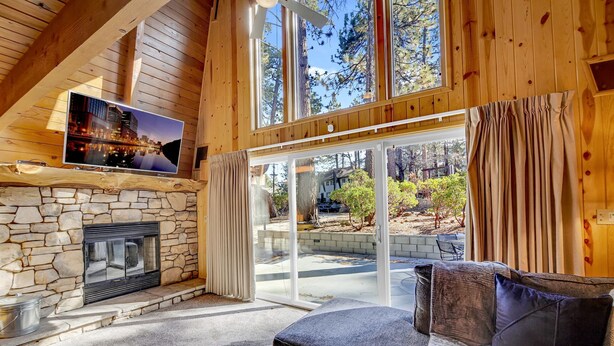 A cozy living room with a stone fireplace, a flat-screen TV mounted on the wall, and a large sliding glass door leading to an outdoor area. - Switzerland SKI Chalet #845 (Big Bear Lake)