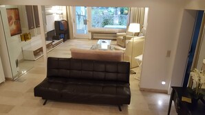 Una sala de estar moderna con un sofá de cuero negro, un sillón beige, una pequeña mesa de centro y un ventanal con vistas al exterior.