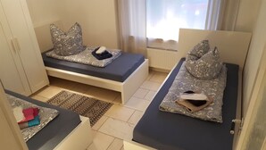Habitación pequeña con dos camas individuales, cada una con almohadas estampadas y un colchón oscuro. Se ve un radiador al fondo, y un tapete con motivos decorativos cubre el suelo.