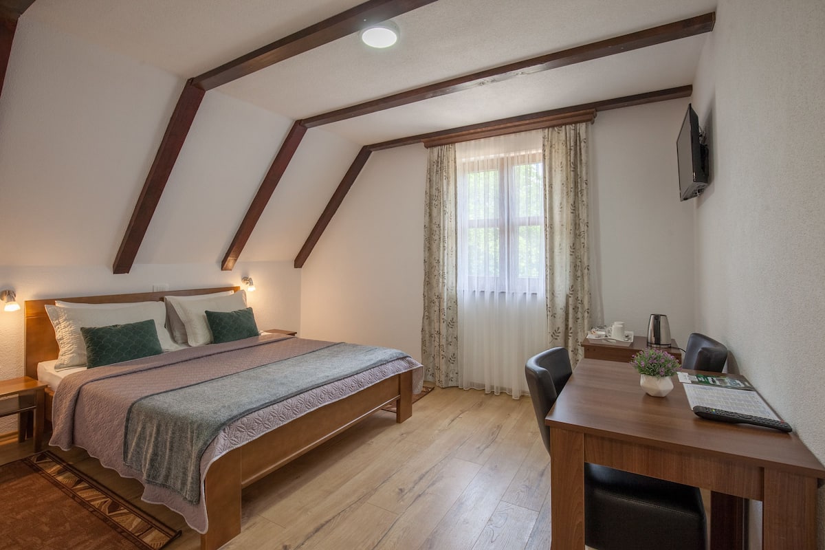 A bedroom with a wooden bed, a desk, and a chair. There is a window with curtains and a TV mounted on the wall.