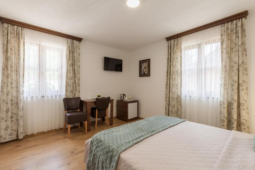 A bedroom with a bed, a desk, two chairs, a small cabinet, a television, and curtains.