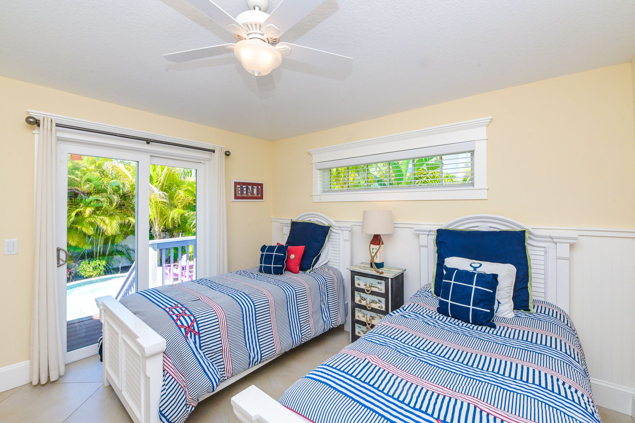 A bedroom with two beds, a ceiling fan, a window with a view of greenery, and a door leading to an outdoor area.