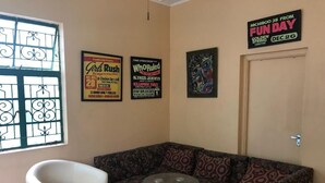 A living room with a white armchair, a floral sofa, and a small round table. There are posters on the wall advertising events and a door with a sign for a 'Fun Day' event. - Manor Park Private Room (Constant Spring)