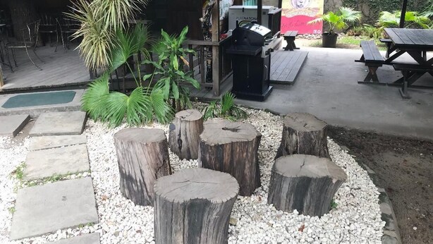An outdoor seating area with wooden tables and benches, surrounded by a garden with plants and a barbecue grill. - Manor Park Private Room (Constant Spring)