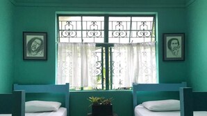 A room with teal walls, two single beds with white bedding, a small table with a plant, and a window with decorative iron bars and sheer curtains. - Manor Park Private Room (Constant Spring)