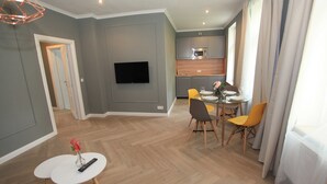 Ein modernes Wohnzimmer mit Flachbildfernseher, Essbereich mit kleinem Tisch und Stühlen sowie eine Küche mit grauen Schränken und einer Mikrowelle.