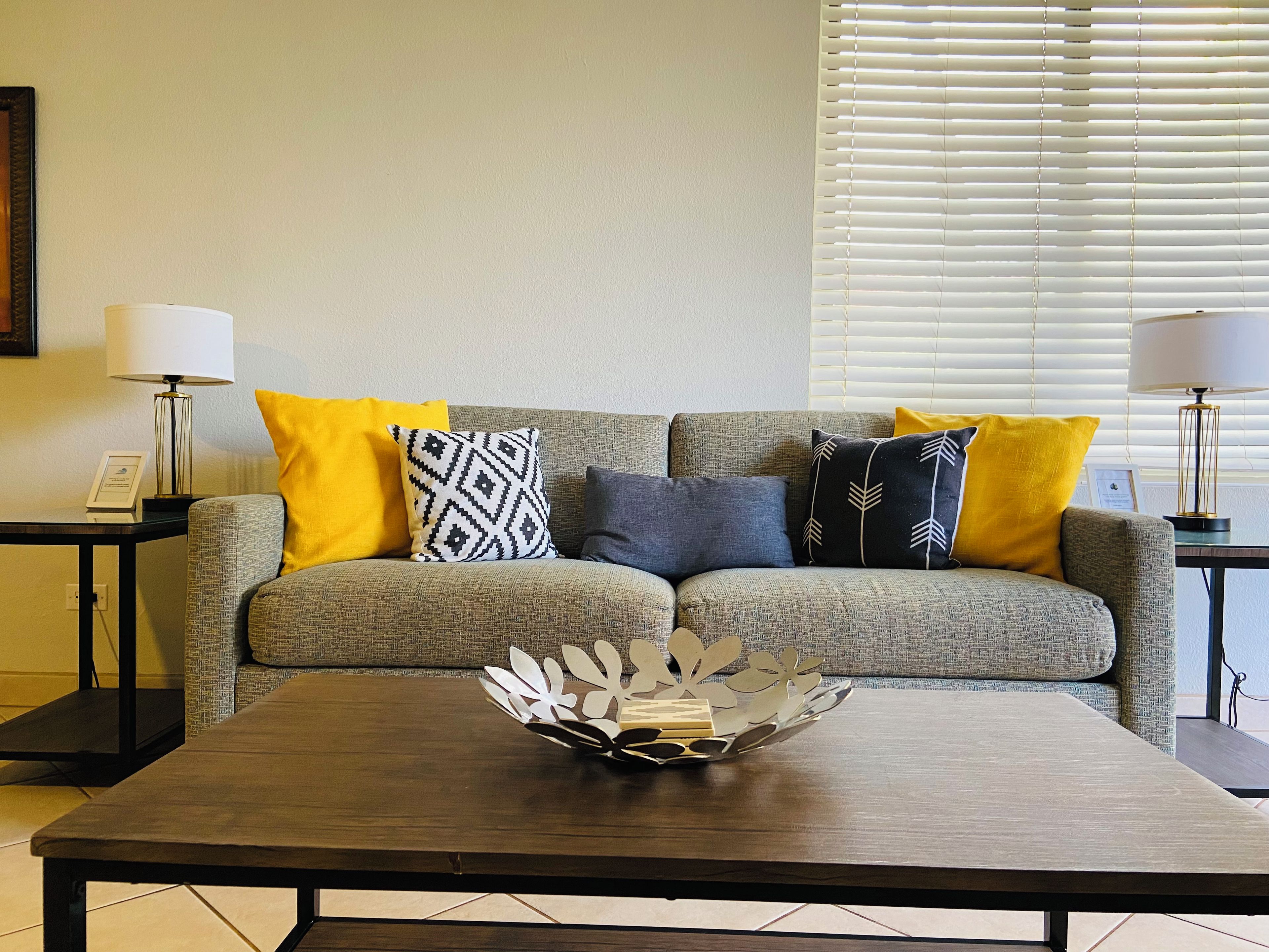 A living room with a grey sofa, yellow and patterned cushions, a wooden coffee table, and two lamps on side tables.