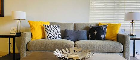 A living room with a grey sofa, yellow and patterned cushions, a wooden coffee table, and two lamps on side tables.