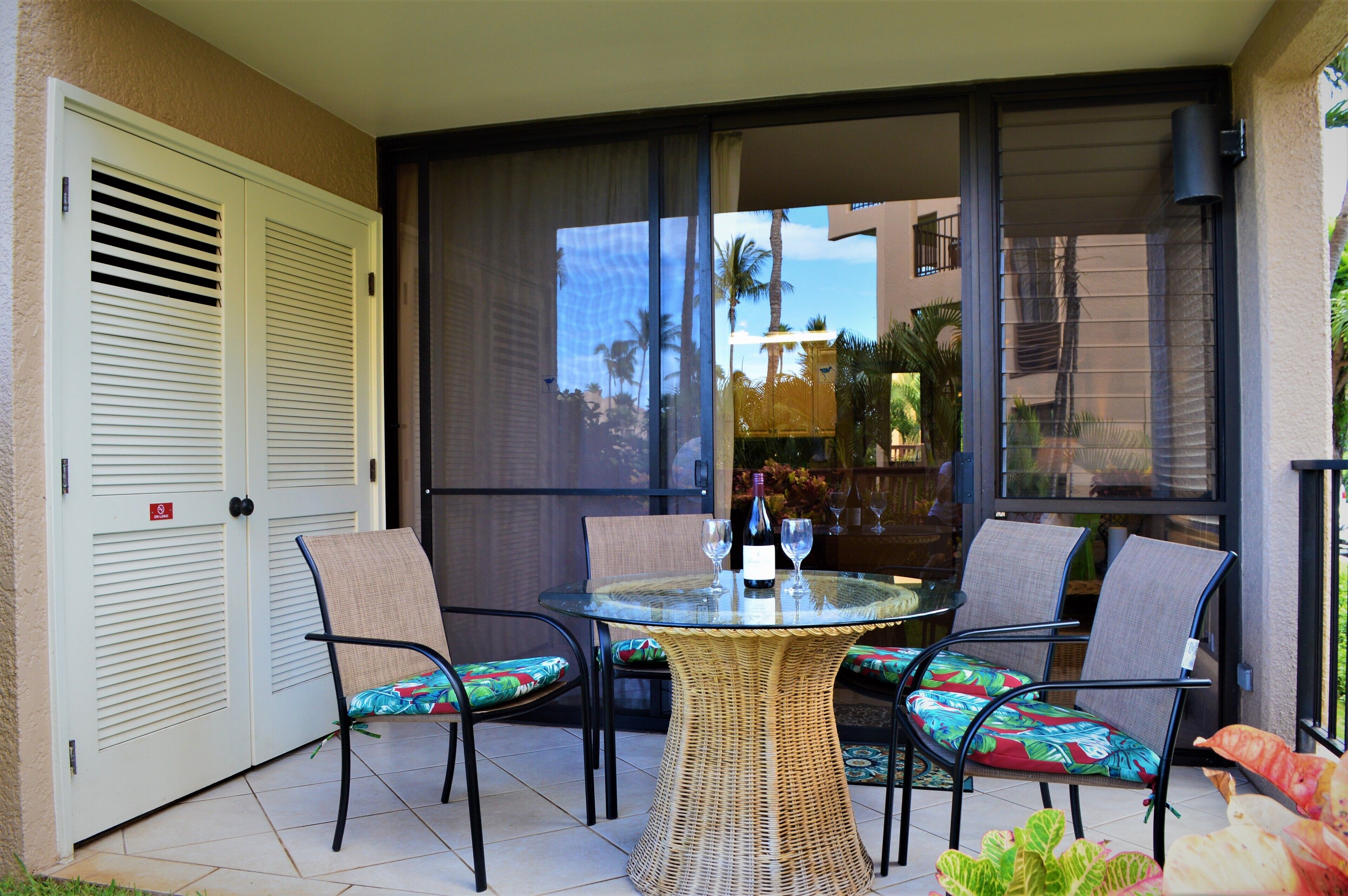 A glass-enclosed patio with a wicker table and four chairs, a sliding glass door, and a view of palm trees and buildings.