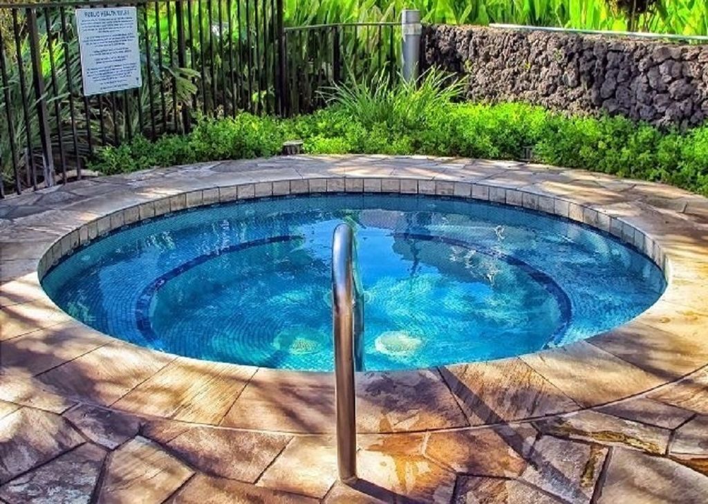 A circular pool with a metal handrail, surrounded by stone tiles and lush greenery.