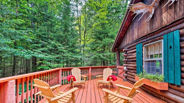 Una terraza de madera con sillas Adirondack, una cabaña con contraventanas verdes y un bosque al fondo.