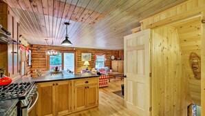 Interior de una cabaña de madera con cocina, que incluye estufa, armarios y comedor con mesa y sillas.