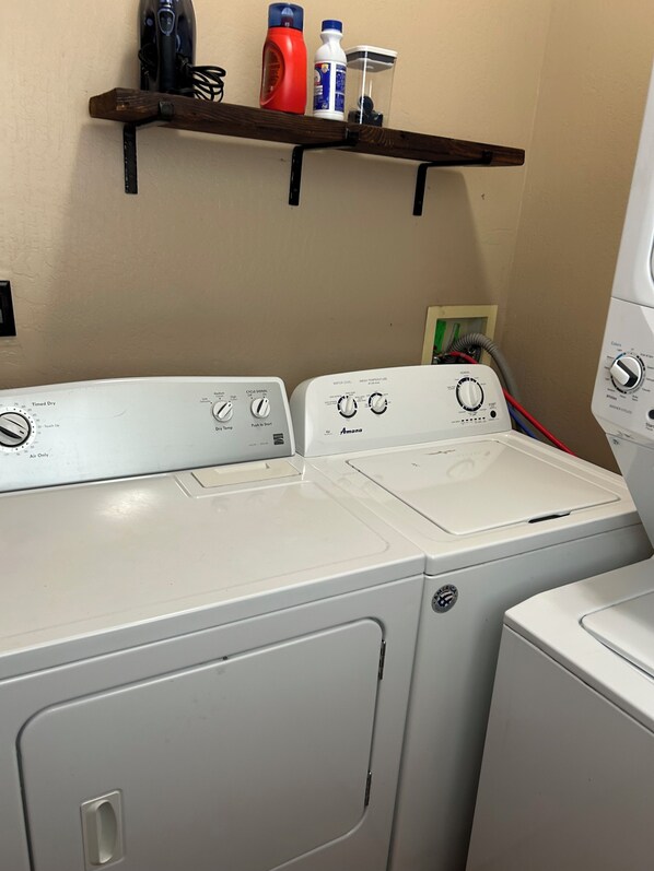 A laundry area with two front-loading washers, a shelf above containing cleaning supplies, and a power outlet with a green switch.